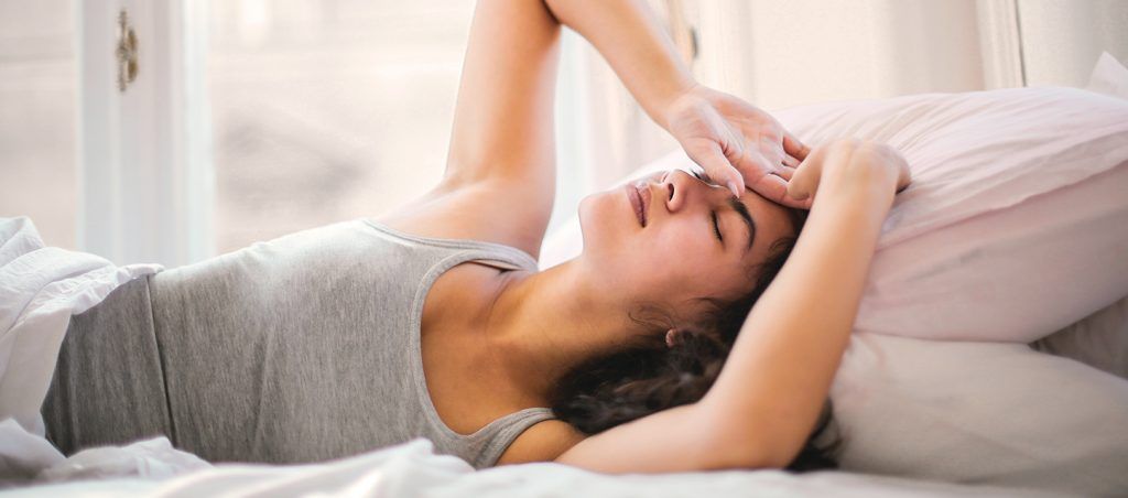 chica de media melena morena tapada dentro de la cama tapada con edredon hasta la cintura boca arriba con las manos en la frente con gesto de malestar por haber dormido en un mal colchon