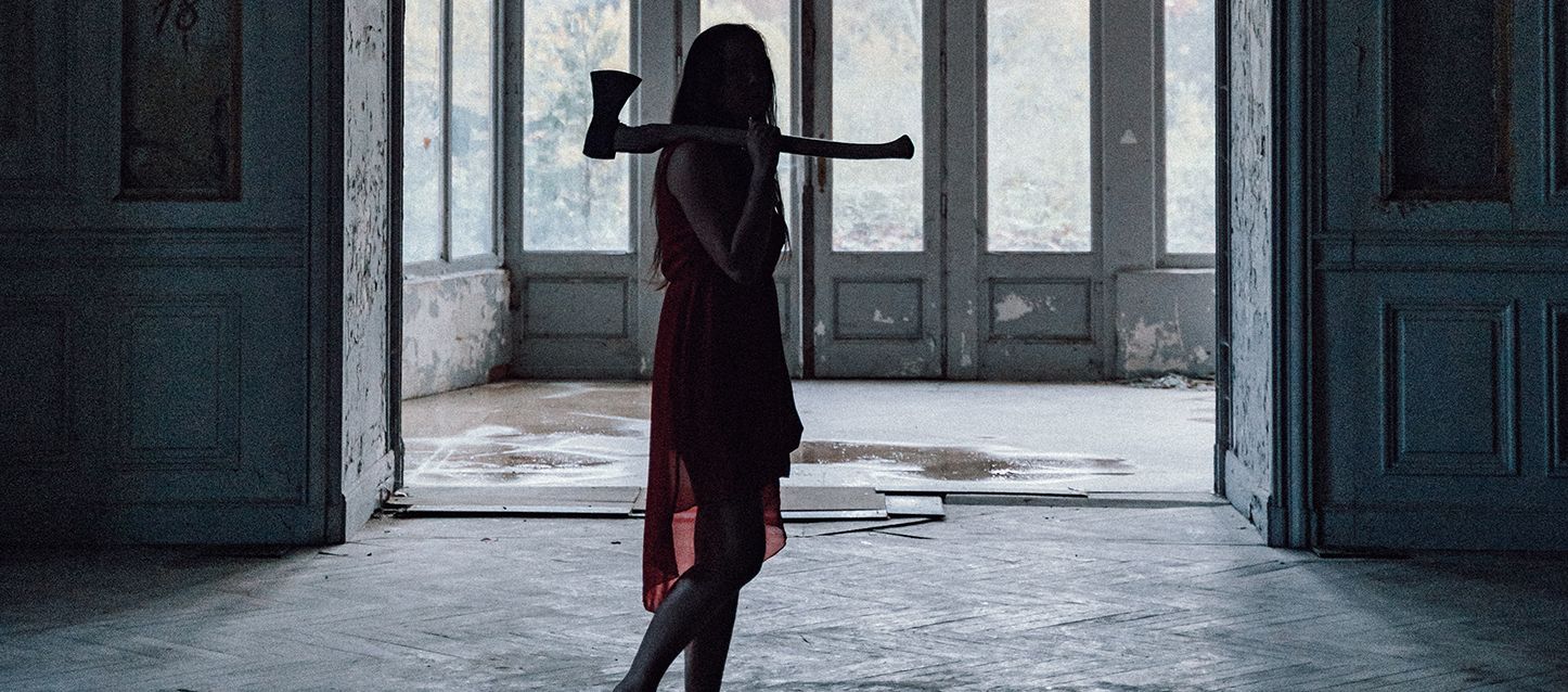 mujer de pelo largo negro de pie con vestido rojo y un hacha al hombro en una casa abandonada