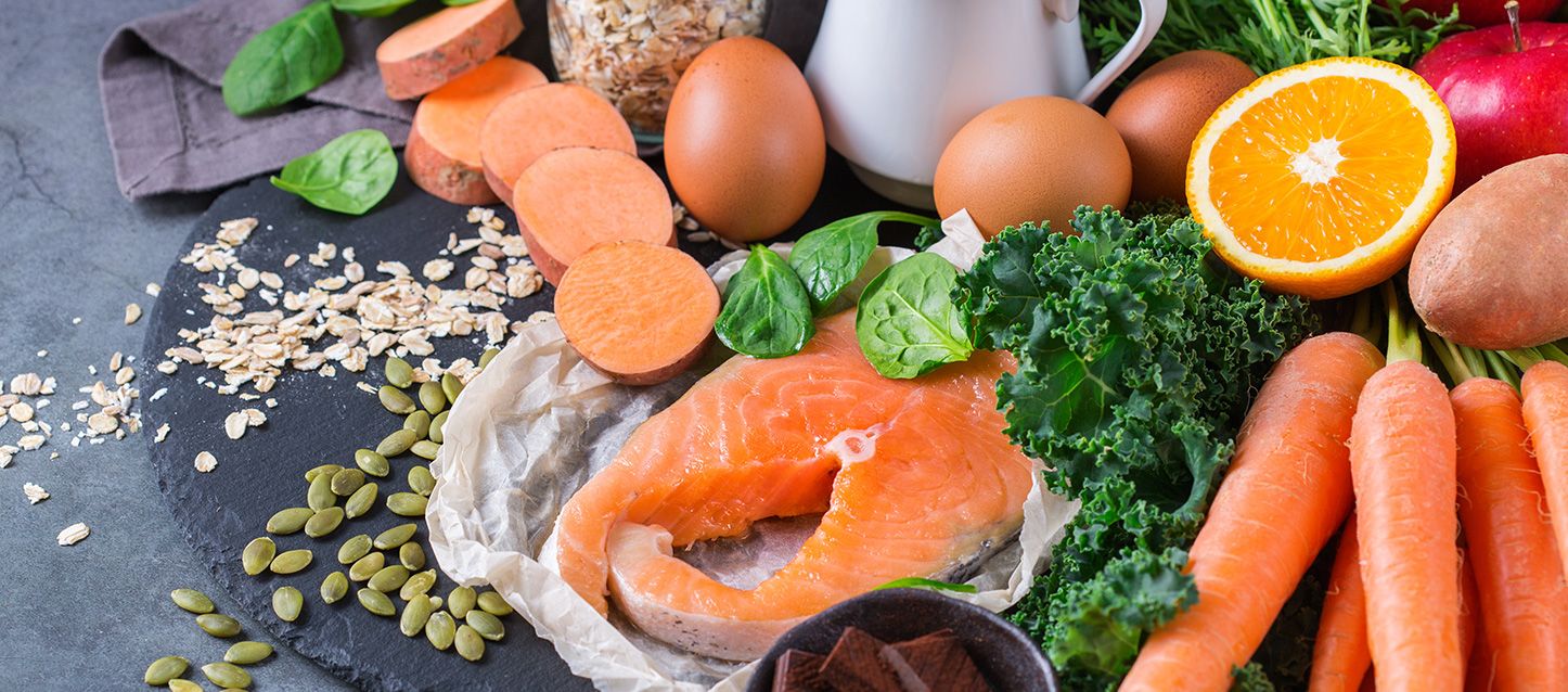 alimentos ricos en vitamina D sobre un plato llano de pizarra como boniatos, salmon, zanahorias, naranja, kale, huevos