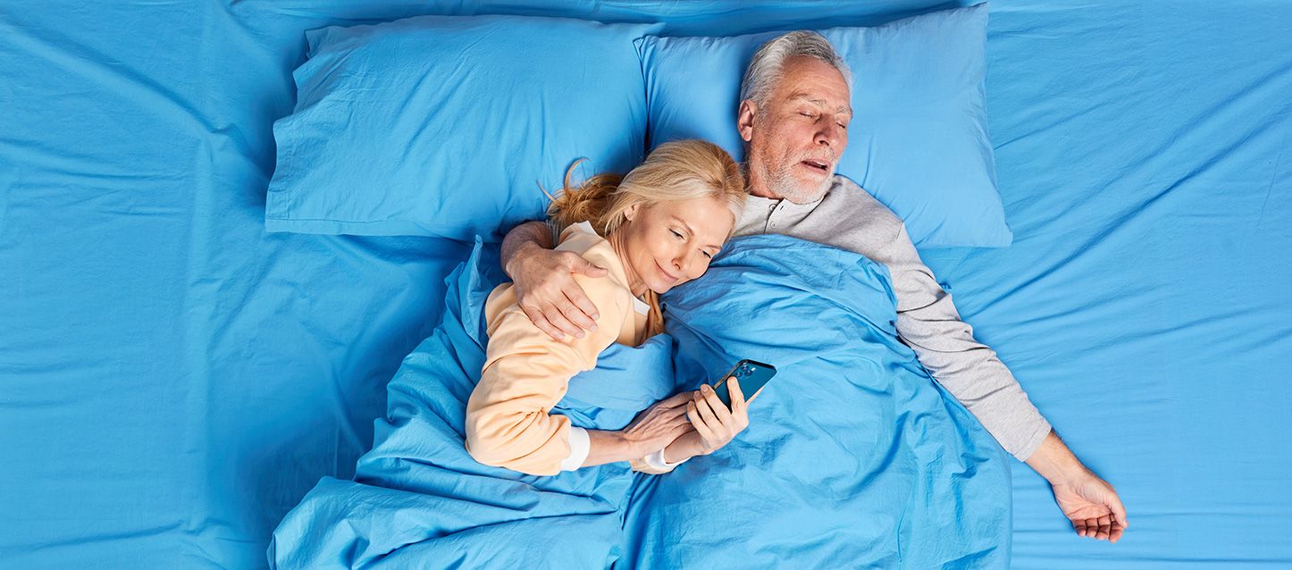 dos personas mayores metidas en la cama donde ella está mirando el móvil y el está durmiendo tapados con edredón y sábanas azules