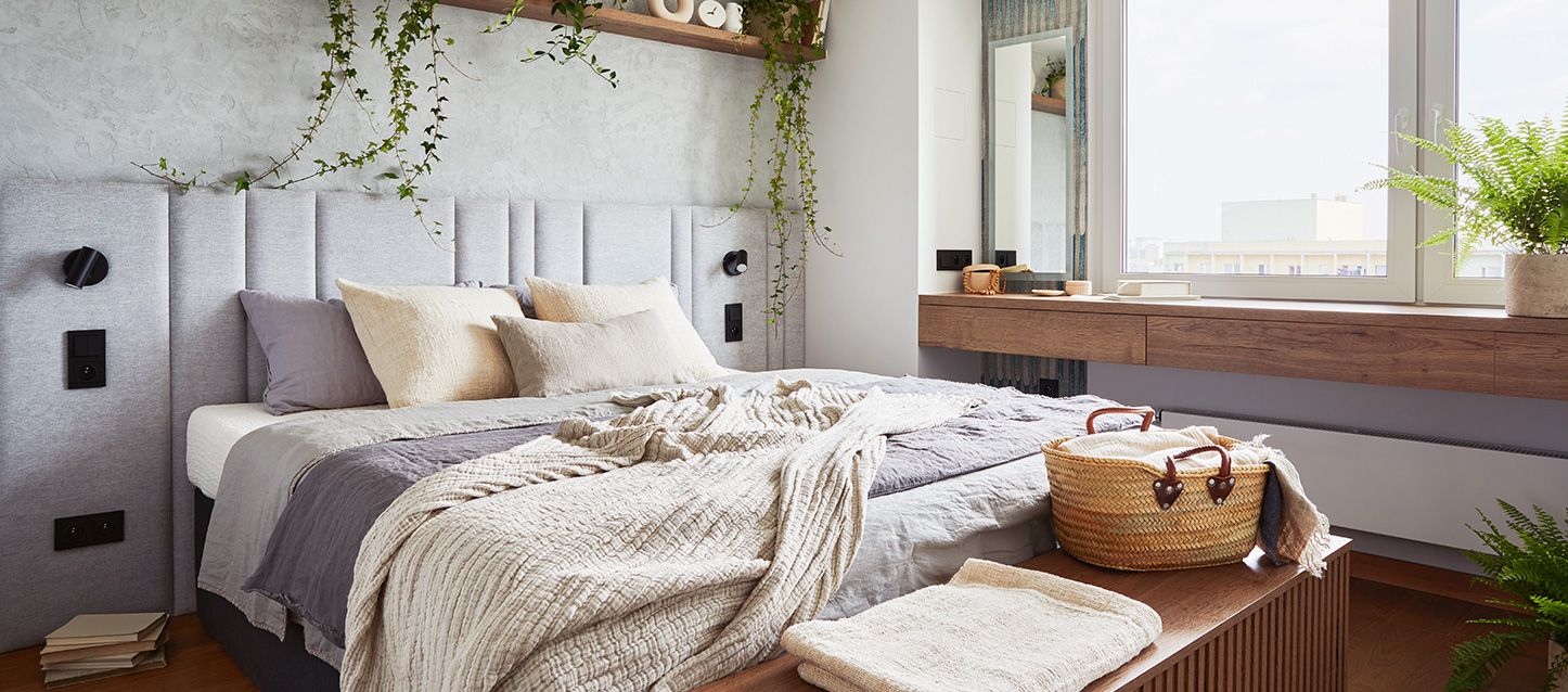 dormitorio de un apartamento pequeño con cama, edredon morado y beige, cojines a juego, cabecero gris y estanteria con objetos y plantas