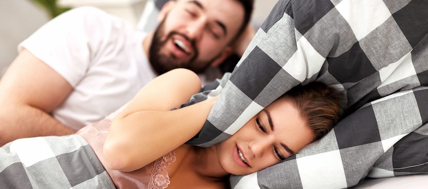 pareja metida en la cama donde el ronca y ella se tapa los oídos con la almohada con gesto de malestar