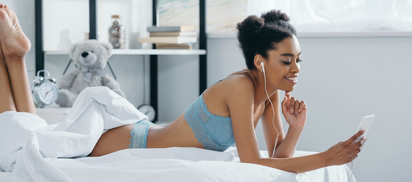 chica de color con el pelo afro tumbada boca abajo en la cama con encaje de lenceria azul apoyada sobre sus codos mirando el móvil y con los auriculares puestos
