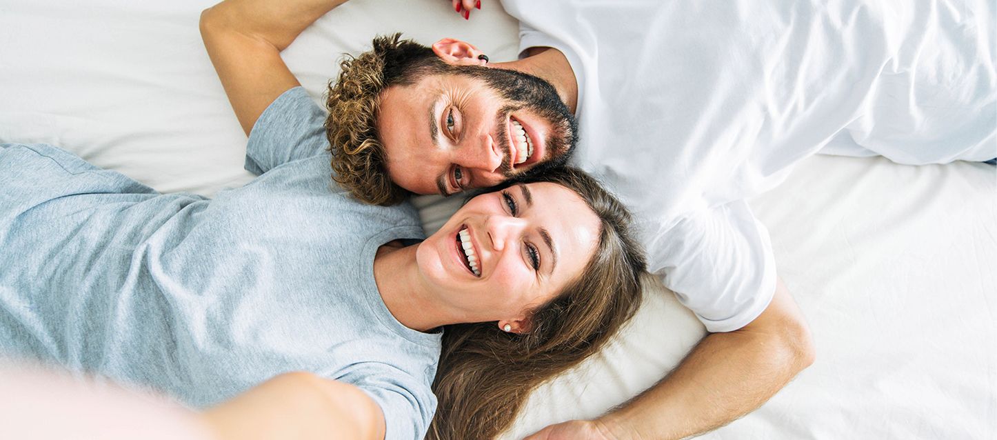 Un chico y una chica sonrientes tumbados boca arriba en sentido opuesto en una cama pero con la cara a la misma altura