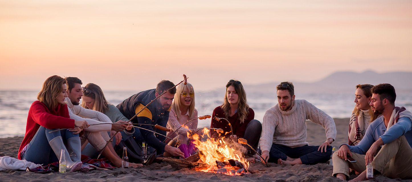 9 Rituales sencillos para la noche de San Juan Un griuo de amigos sentado en la playa junto al mar alrededor de una hoguera asando salchichas al amanecer